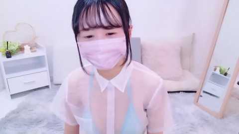 Media: Video of an East Asian woman with straight black hair, wearing a sheer white blouse, revealing a light blue bra underneath, and a pink surgical mask, in a minimalist bedroom with white walls and a beige chair.