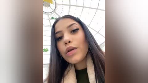 Media: A close-up video of a young woman with long, straight dark hair, wearing dramatic eyeliner and a green top, standing under a white, circular skylight in a modern, indoor setting.