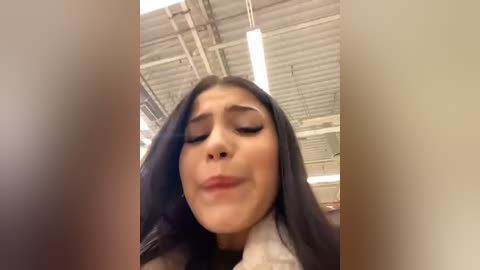 Media: A low-angle, close-up video of a young woman with long, dark hair, wearing makeup, in a classroom with a metallic ceiling and fluorescent lights.