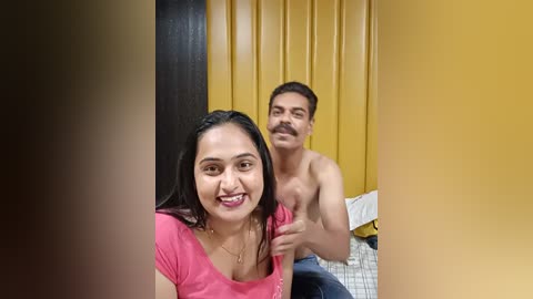 Media: Video of a smiling South Asian woman in a pink top, seated next to a topless man with a mustache, in a room with mustard-yellow curtains and a white bed.