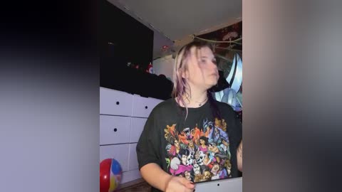 Media: A video of a young woman with long, blonde hair wearing a colorful graphic tee, standing in a cluttered bedroom with white drawers, a colorful beach ball, and a partially visible window.