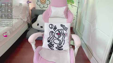 Media: A video of a pastel pink gaming chair with a white pillow featuring a cartoonish, smiling face design. The background includes a white wall, a gray couch with a plush white bear, and a green plant.