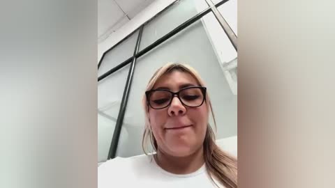 Media: Video of a blonde woman with glasses, fair skin, and a neutral expression, wearing a white shirt, standing in a modern office with large glass windows and white walls.