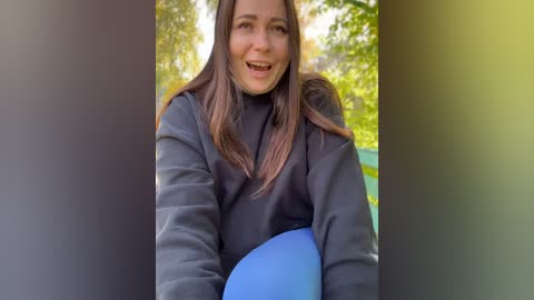 Media: Video of a smiling woman with straight brown hair, wearing a black hoodie, holding a blue object. Background features blurred green foliage.