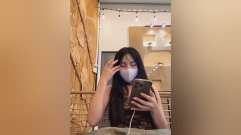 Media: Video of a young woman with long black hair, wearing a face mask, taking a selfie in a cozy caf\u00e9. Background features wooden walls, string lights, and a rustic interior.