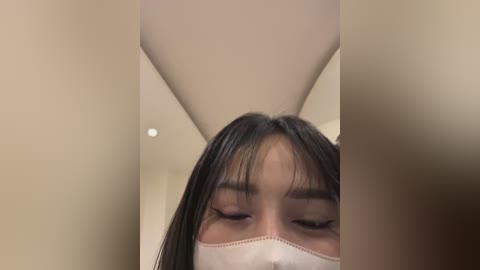 Media: A close-up video of a young Asian woman with straight, black hair, wearing a white surgical mask, taken from a low angle, showcasing her closed eyes and serene expression. The background features a beige wall and a ceiling light.