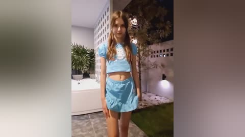 Media: Video of a young, slender, light-skinned girl with long blonde hair wearing a blue crop top and shorts, standing in a modern, well-lit backyard with a jacuzzi, potted plants, and a white fence.