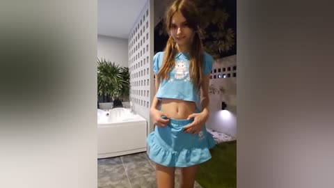 Media: Video of a young, slender Caucasian girl with long brown hair in a blue, short-sleeved, ruffled crop top and matching skirt, standing indoors near a white bathtub, potted plants, and decorative wall panels.