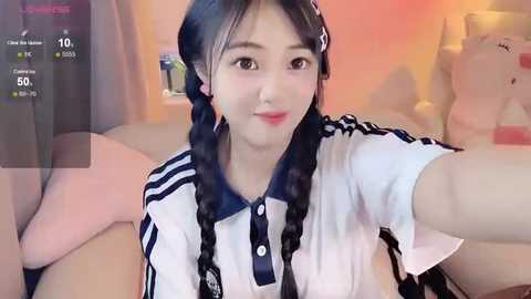 Media: A video of an East Asian woman with long black hair in twin braids, wearing a sailor-style school uniform, smiling and taking a selfie in a cozy, warmly-lit room.