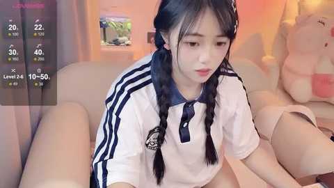 Media: Video of an Asian woman with long black pigtails, wearing a white Adidas shirt, sitting on a beige couch. Background includes a plush toy, a fish tank, and a warm orange light.