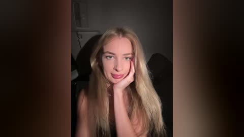 Media: Video of a blonde woman with fair skin and green eyes, resting her chin on her hand, smiling softly. She wears a black top, set against a dimly lit, indistinct background.