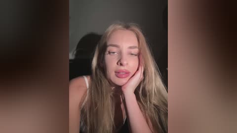 Media: Video of a young woman with long, wavy blonde hair, fair skin, and pink lips, resting her head on her hand, wearing a black tank top, in a dimly lit room.