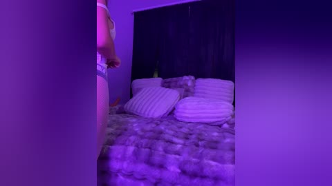 Media: Video of a dimly-lit bedroom with purple lighting. A person in a white tank top is seen partially through a doorway, standing next to a bed with white pillows and a patterned blanket.