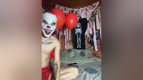 Media: Video of a shirtless, pale-skinned man with a clown mask, red hair, and tattoos, holding a red balloon. Background features a skeleton, a bed, and festive decorations.