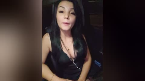 Media: A video of a Latina woman with long black hair, medium complexion, wearing a black sleeveless top, sitting in a dark room. She has a thin build and medium-sized breasts.