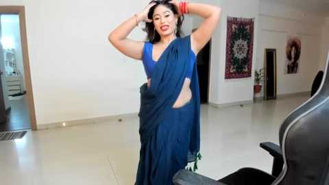 Media: Video of a South Asian woman in a blue saree, striking a pose in a modern, minimalist room with beige floors, red tapestry, and a black gaming chair.