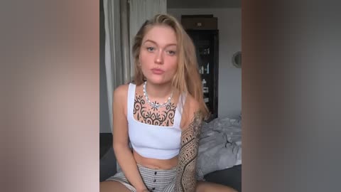 Media: Video of a young woman with long blonde hair, wearing a white crop top revealing intricate tattoos, sitting on a bed with grey sheets in a dimly lit bedroom.