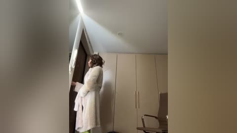 Media: A video of a woman in a white robe standing in a modern, minimalist bedroom with beige walls and a large, white wardrobe. She is looking out a window, bathed in soft sunlight.