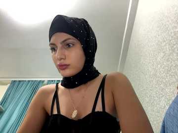 Media: Video of a woman with medium skin tone, wearing a black headscarf, black spaghetti-strap top, and gold necklace, in a room with a textured wall and teal curtain.