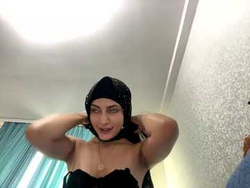 Media: Video of a woman with light skin, wearing a black hijab and a strapless black top, posing confidently with hands on head in a room with teal curtains and textured white walls.