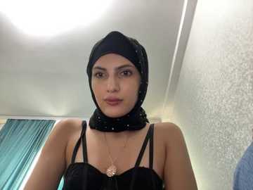 Media: Video of a young woman with olive skin and dark hair under a black hijab, wearing a black top with thin straps, standing indoors with a textured wall and teal curtains in the background.