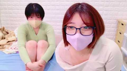 Media: Video of two East Asian women, one with short black hair, wearing a green cardigan and beige mask, sitting on a bed. The other, with shoulder-length red hair, glasses, and purple mask, sits on the bed. White brick wall background.