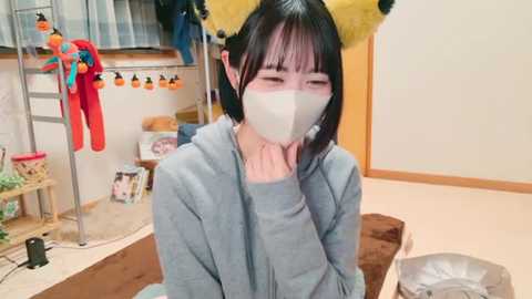 Media: Video of an Asian woman with short black hair, wearing a gray hoodie and a white face mask, seated in a cozy, cluttered room with a shelf full of plush toys, a brown sofa, and a small table.