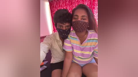 Media: Video of two young South Asian girls with dark skin, wearing face masks and colorful striped shirts, sitting in a room with red curtains and a pinkish background.