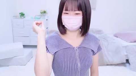 Media: A video of an East Asian woman with straight, shoulder-length black hair, wearing a light purple striped top and a white face mask, standing in a minimalist, white bedroom with a bed and dresser in the background.