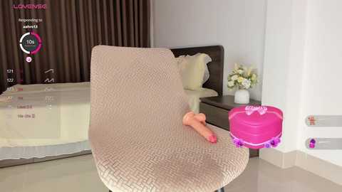 Media: Video of a modern bedroom with beige carpet, a pink heart-shaped vibrator on a bed, and a floral arrangement on a nightstand.