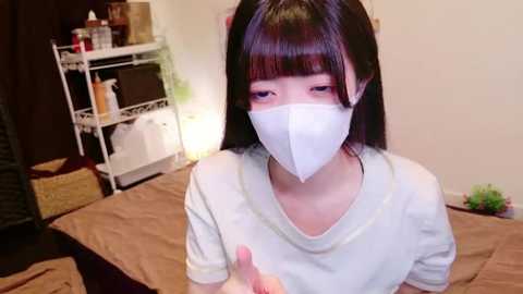 Media: Video of a young Asian woman with straight black hair, wearing a white mask and light-colored shirt, seated on a bed in a cozy, dimly lit room with a white shelf holding household items in the background.