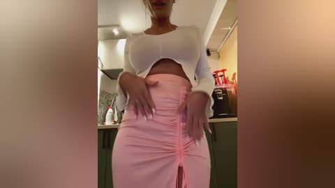 Media: Video of a light-skinned woman in a tight, white crop top and high-waisted, pink pleated skirt, standing in a modern kitchen with stainless steel appliances and red coffee maker.