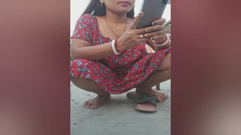 Media: Video of a South Asian woman squatting, holding a smartphone, wearing a red floral dress, brown sandals, and bracelets, in a concrete courtyard.