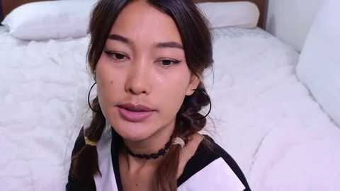 Media: Video of a young woman with light brown skin and dark hair in pigtails, wearing a white and black jacket, black choker, and makeup, sitting on a white bed with a brown headboard.