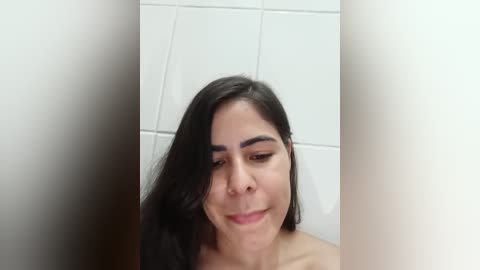 Media: Video of a Latina woman with long black hair and medium skin tone, smiling slightly, in a white-tiled bathroom. She has dark eyebrows and is topless.