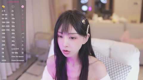 Media: A video of an East Asian woman with long black hair, wearing a white off-shoulder dress, in a modern living room.