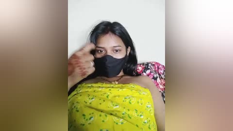 Media: Video of a woman with medium-dark skin, wearing a black mask, floral-patterned yellow dress, and floral-patterned sleeveless top. She raises her right hand near her face, partially obscuring it.