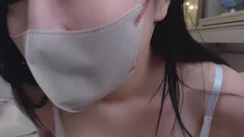 Media: A close-up video of a woman with fair skin, wearing a white surgical mask and a blue bra strap. Her long black hair is slightly damp, and she is indoors near a mirror and a cluttered shelf.