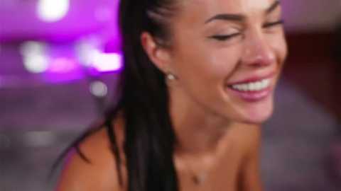 Media: A close-up video of a smiling, topless woman with wet, slicked-back black hair and earrings. Background is blurred with purple and white lights.