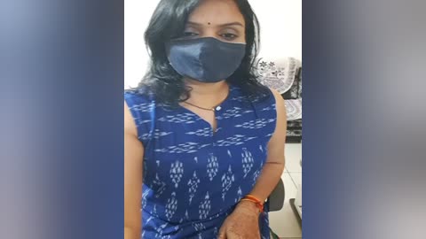 Media: Video of a South Asian woman with medium skin tone, wearing a blue patterned blouse, black face mask, and a red bangle. She stands in an indoor setting with a white wall and decorative items in the background.