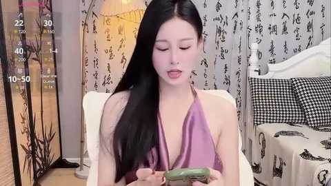 Media: Video of an Asian woman with long black hair, wearing a purple halter top, seated on a bed, looking at a green phone, surrounded by patterned curtains and a checkered pillow.