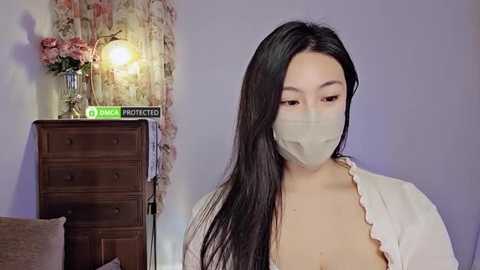 Media: Video of an Asian woman with long black hair, wearing a beige mask, white ruffled top, and a floral-patterned backdrop, holding a book.