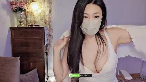 Media: Video of an Asian woman with long black hair, wearing a white lace bralette, gray face mask, and a gray wig, standing in a bedroom with a wooden dresser, floral arrangement, and white bedding.