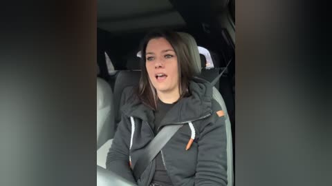 Media: Video of a woman with straight brown hair, wearing a gray hoodie and seatbelt, seated in a car, mouth open in apparent surprise or excitement.