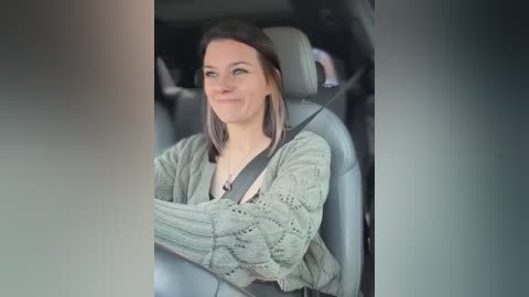 Media: Video of a smiling woman with shoulder-length brown hair, wearing a light green knitted cardigan, driving in a car with a grey seatbelt and dark interior.