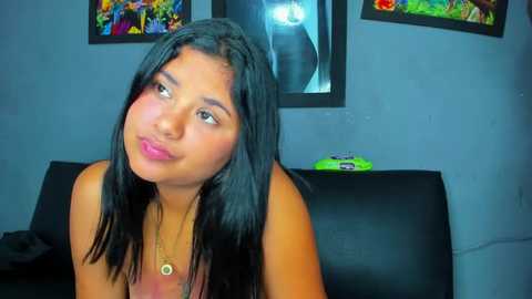 Media: Video of a young woman with straight black hair, light skin, and a small build, wearing a gold necklace. She sits on a black couch in a dimly lit room with colorful paintings on the wall.