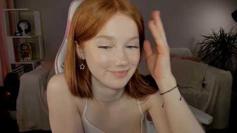 Media: Video of a fair-skinned, red-haired young woman with freckles, wearing a striped headband and star earrings, smiling while playing with her hair, set in a cozy living room with shelves and potted plants.