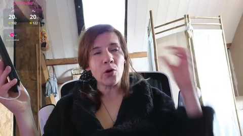 Media: A candid video of an Asian woman with straight brown hair, wearing a black fur coat, sitting in a dimly lit room. She holds a smartphone, appearing to record herself.