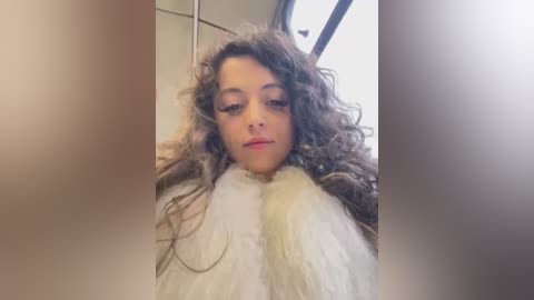 Media: Video of a young woman with curly brown hair, wearing a fluffy white robe, sitting in a train. She has a neutral expression, and the background shows train windows and interior.