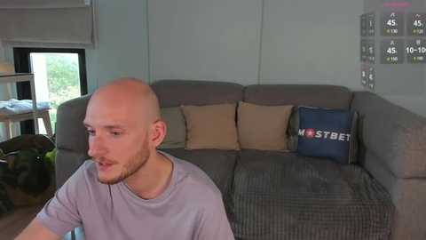 Media: Video of a bald, bearded man in a gray shirt, sitting on a gray couch with beige and blue cushions, in a modern living room with white walls, a window, and a sports score board.